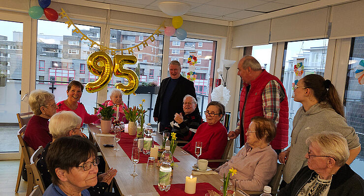 Eine Gruppe Menschen steht und sitzt um eine Kaffeetafel herum.