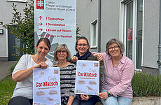 Vier Frauen sitzen nebeneinander und halten Plakate mit der Aufschrift Café Cariklatsch hoch.