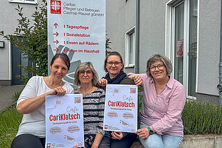Vier Frauen sitzen nebeneinander und halten Plakate mit der Aufschrift Café Cariklatsch hoch.