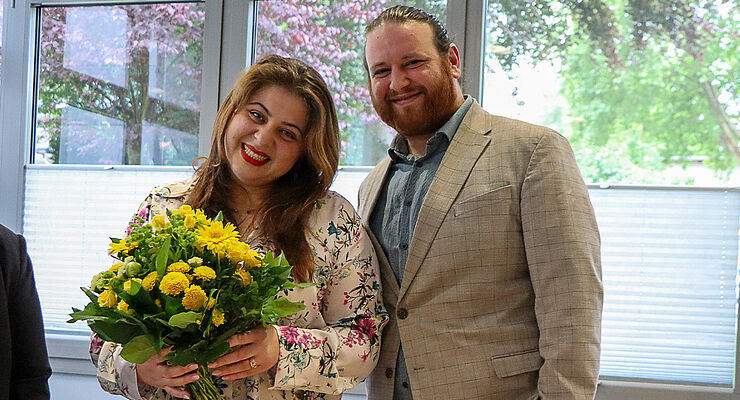 Eine Frau mit Blumen und ein Mann stehen nebeneinander und lächeln.