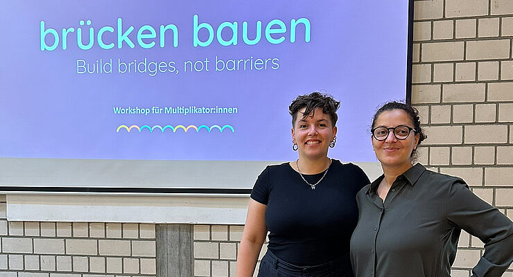 Zwei Frauen stehen vor einer Leinwand mit der Aufschrift "brücken bauen"