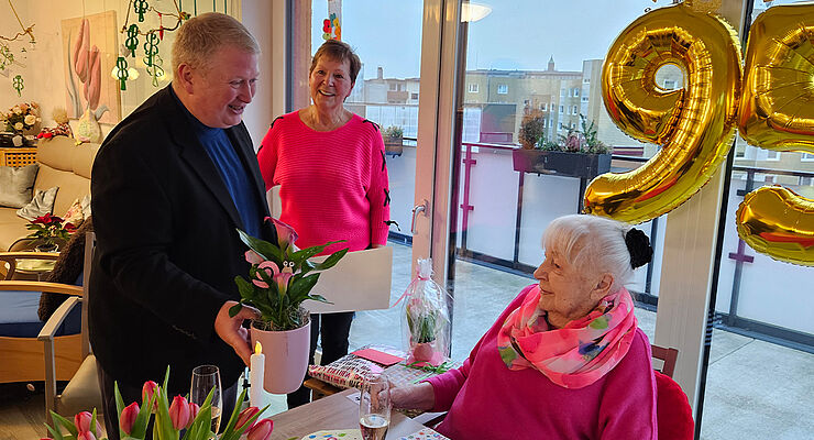 Ein Mann überreicht einer älteren Dame Blumen, im Hintergrund lächelt eine Frau.