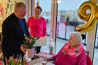Ein Mann überreicht einer älteren Dame Blumen, im Hintergrund lächelt eine Frau.