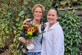 Zwei Frauen stehen nebeneinander, eine Frau hält Blumen in den Händen.