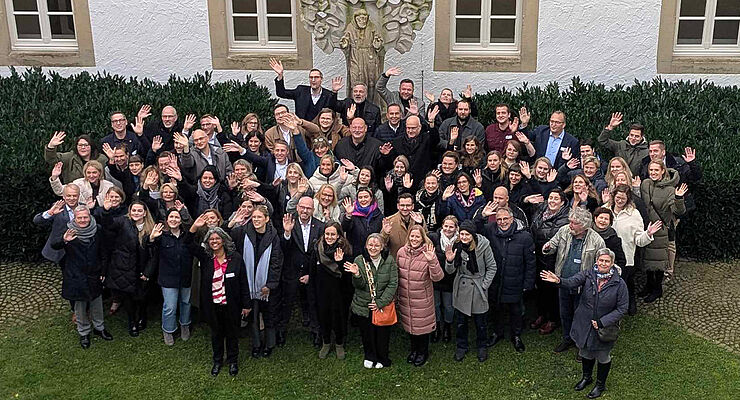 Eine große Gruppe Menschen steht auf einer Wiese zusammen und winkt.